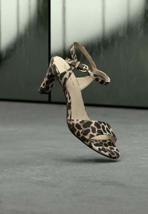 Leopard-print high-heeled sandal with ankle strap and gold buckle, positioned above a gray floor with a blurred green background.