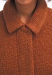 Brun texturerad jacka med ett tjockt, mjukt och fluffigt tyg. Har en bred krage och en knappslå framtill. Materialet har ett mjukt, öglat utseende.