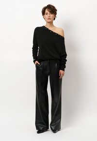 Schwarzer Off-Shoulder-Pullover mit silbernen Nieten entlang des Ausschnitts, kombiniert mit hoch taillierten schwarzen Leder-Hosen mit weitem Bein.