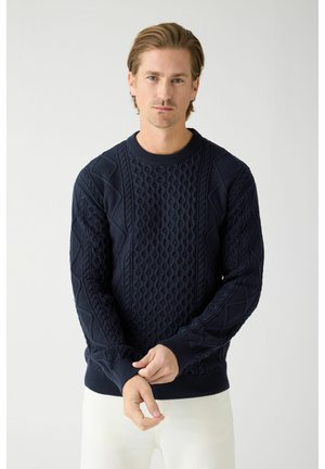 Mand iført en mørk marineblå kabelstrikket sweater og hvide bukser, justerer en ærme med et neutralt udtryk mod en lys baggrund.