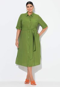 Abito verde con bottoni, maniche corte, cintura in vita e aperture laterali. La modella indossa sandali arancioni con cinturini. Tessuto liscio.