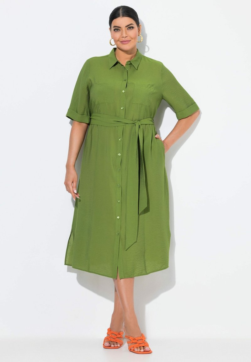 Abito verde con bottoni, maniche corte, cintura in vita e aperture laterali. La modella indossa sandali arancioni con cinturini. Tessuto liscio.