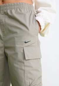 Svetlo sivi delovni hlače z elastičnim pasom, sprednjimi žepi in črnim Nike logotipom na stegnu, ki prikazujejo gladko, teksturirano tkanino.