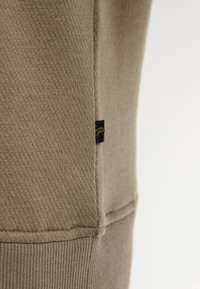 Sudadera beige con textura sutil, que presenta una costura lateral y una pequeña etiqueta negra con una "P" naranja. Tejido suave y dobladillo acanalado.