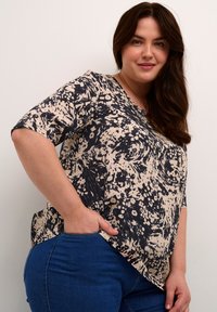 Haut à manches courtes beige avec un motif abstrait noir, doté d'un col en V et d'une coupe décontractée, associé à un jean en denim bleu.