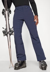 Ναυτικές παντελόνες χιονιού με φερμουάρ, κατασκευασμένες από αδιάβροχο υλικό. Έχουν χαλαρή εφαρμογή και είναι συνδυασμένες με μαύρες μπότες σκι και σκι.