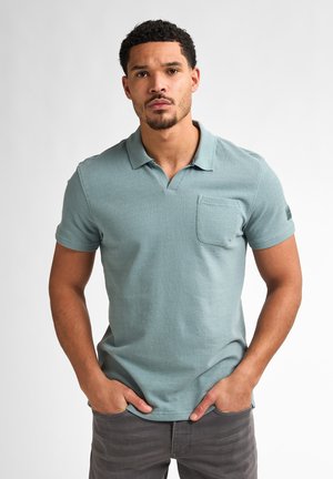 Camisa polo en verde azulado apagado, de mangas cortas, hecha de tejido texturizado, con un bolsillo en el pecho y un sutil detalle del logo en la manga.