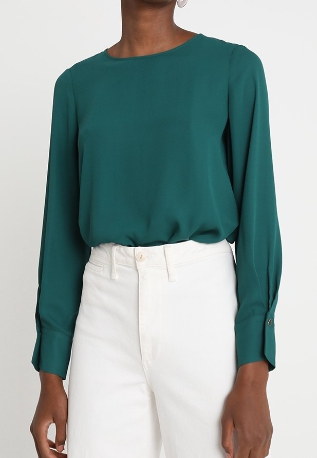 Blusa de manga larga en teal de tejido ligero con cuello redondo y puños abotonados, combinada con pantalones blancos de talle alto.