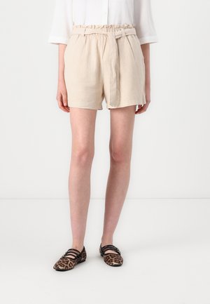 Persona que lleva pantalones cortos beige con cinturón estilo paperbag en la cintura, combinados con zapatos planos con estampado de leopardo y tiras negras, de pie sobre un fondo blanco.