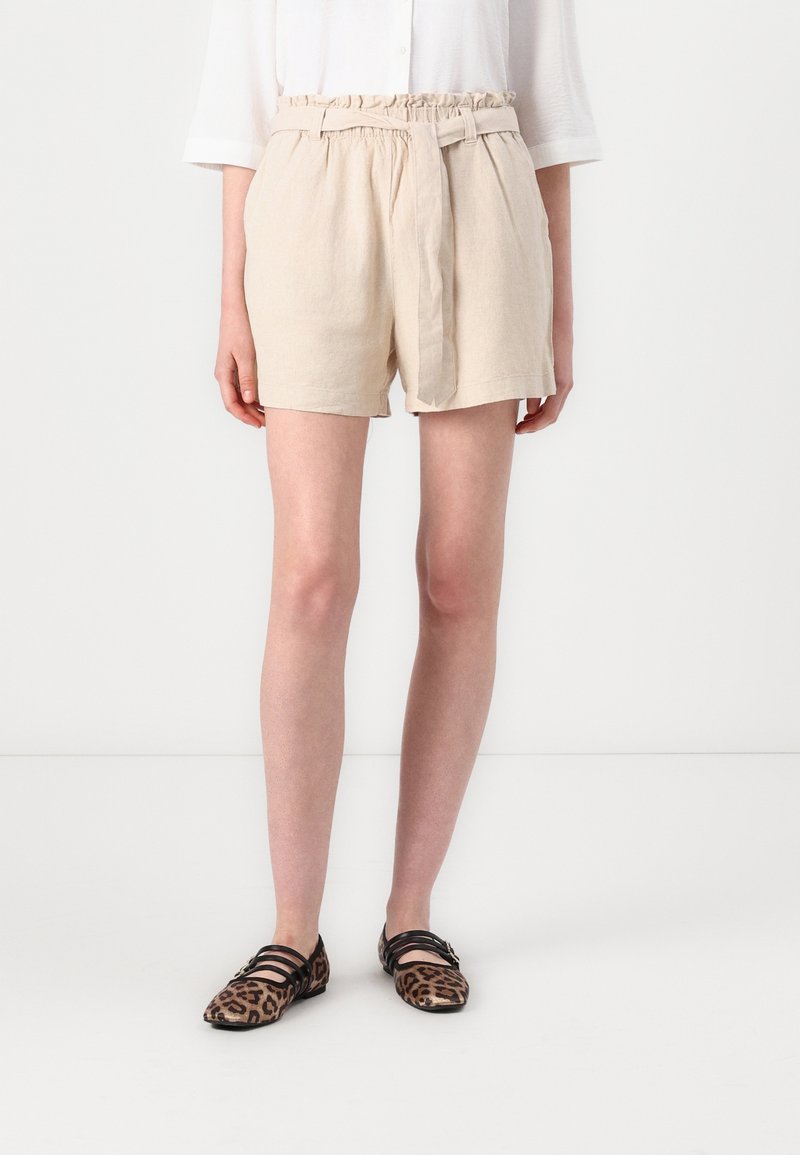Personne portant un short beige taille paperbag ceinturé, associé à des ballerines imprimé léopard avec des lanières noires, debout sur un fond blanc.