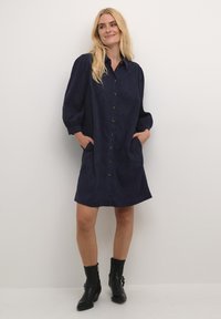 Robe chemise bleu marine en matériau doux, dotée d'un devant boutonné, de deux poches latérales et de manches ballon. Portée avec des bottines noires.