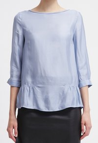 Ljusblå blus med trekvartslånga ärmar, peplumslut och jämn textur. Har rund halsringning och en subtil lyster.