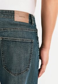 Ciemnoniebieskie dżinsy z denimu z naszywką ze skóry na pasku oznaczoną napisem "DENIM SUPPLY". Posiadają klasyczne tylne kieszenie i prostą nogawkę.