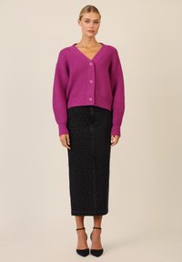 Paarse gebreide cardigan met een V-hals, geribbelde textuur en drie knopen, gecombineerd met een zwarte spijkerrok met zilveren stiksels.