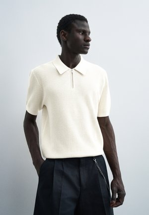 Mand iført en hvid kortærmet strikket polo med lynlås, kombineret med sorte plisserede bukser og en kædeaccessory, kigger til siden.