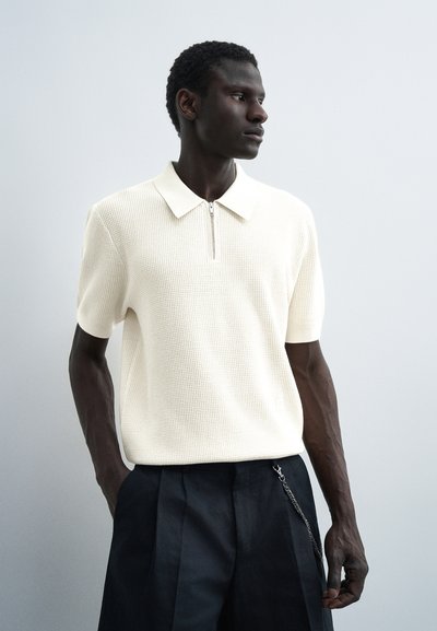 Homme portant un polo en maille blanche à manches courtes avec une fermeture éclair, associé à un pantalon plissé noir et un accessoire chaîne, regardant sur le côté.