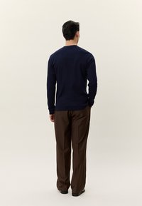 Egy férfi háttal áll, sötétkék hosszú ujjú ingben, barna nadrágban és fekete cipőben egy egyszerű, világos háttér előtt.