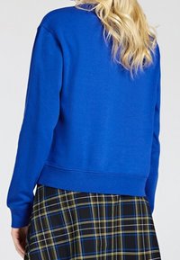 Persona rubia con una blusa de manga larga azul brillante y una falda de cuadros negra, azul y amarilla, vista desde atrás.