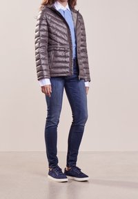 Grå vadderad jacka med dragkedja framtill och sidofickor, matchad med blå skjorta och mörkblå jeans. Modellen bär marinblå sneakers.