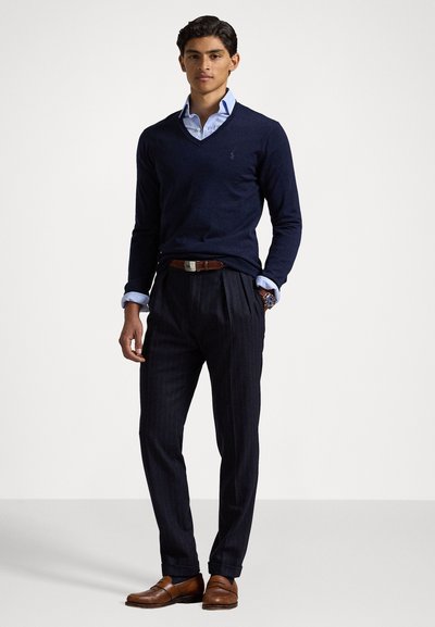 Suéter de pico en color navy sobre una camisa de cuello azul claro, combinado con pantalones oscuros de rayas y zapatos de cuero marrón con un detalle de hebilla.