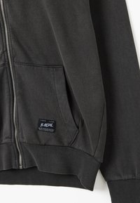 Felpa nera con zip realizzata in tessuto morbido con una finitura testurizzata. Presenta una tasca a marsupio e polsini a costine, con un'etichetta sulla tasca.