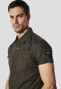 Camisa de manga corta color verde oliva con cierre de botones en el frente, dos bolsillos en el pecho y puños de color azul oscuro contraste. Hecha de tela ligera.