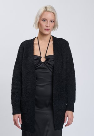 Blonde Frau trägt eine schwarze strukturierte Strickjacke über einem schwarzen Satinkleid mit Neckholder-Ausschnitt und einem runden Detail an der Brust.