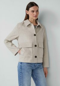 Beige wollen jas met een kraag, een sluiting met knopen aan de voorkant en twee opgestikte zakken. Heeft lange mouwen en een ontspannen pasvorm. Gestreepte top eronder.