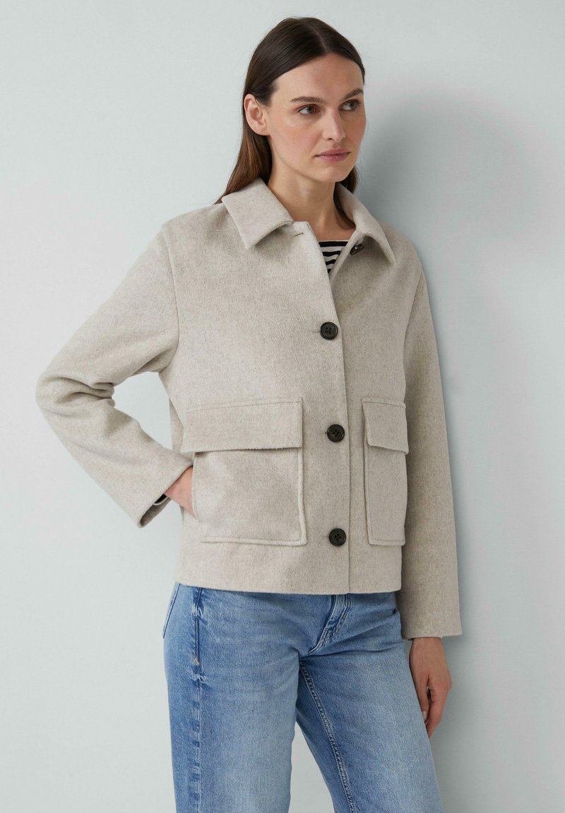 Beige wollen jas met een kraag, een sluiting met knopen aan de voorkant en twee opgestikte zakken. Heeft lange mouwen en een ontspannen pasvorm. Gestreepte top eronder.