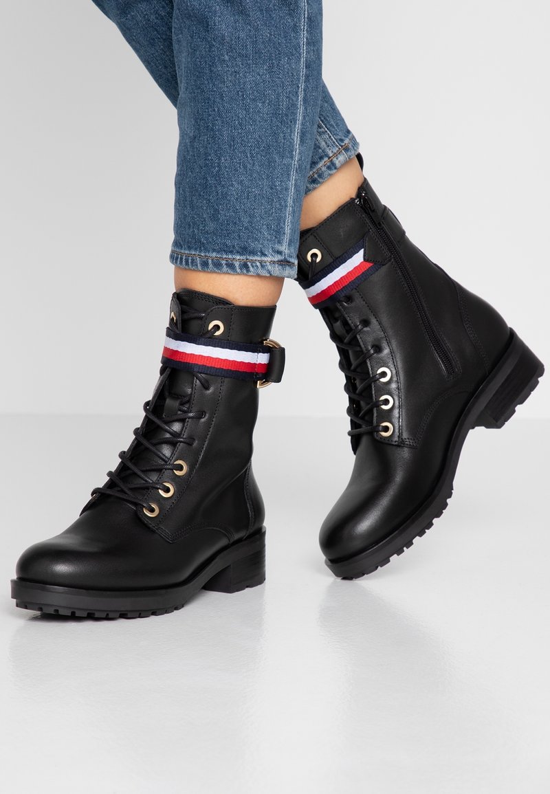 Bottes en cuir noir avec un design à lacets, des œillets dorés, et une bande d'accent en tissu rayé rouge, blanc et bleu autour de la cheville.