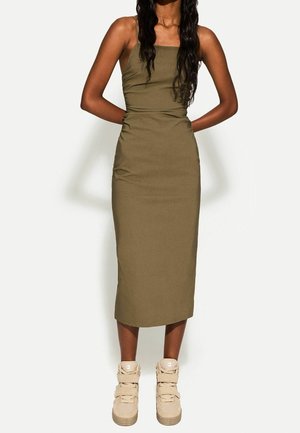 Vrouw in een olijfgroene mouwloze midi-jurk en beige hoge sneakers, staand met haar handen achter haar rug.