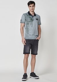 Siva polo majica z črnim ovratnikom, ki ima zelene grafike in besedilo; v kombinaciji z temnimi jeans kratkimi hlačami in črnimi športnimi copati.
