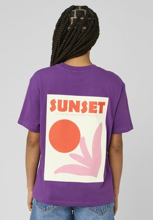 Personne aux cheveux tressés portant un t-shirt violet avec un motif d'un soleil orange et d'une feuille rose avec l'inscription "SUNSET" au dos, sur fond gris.