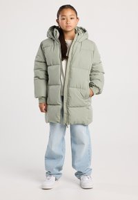 Joven con una chaqueta acolchada con capucha verde claro, jeans azules claros y zapatillas blancas, de pie con las manos en los bolsillos.