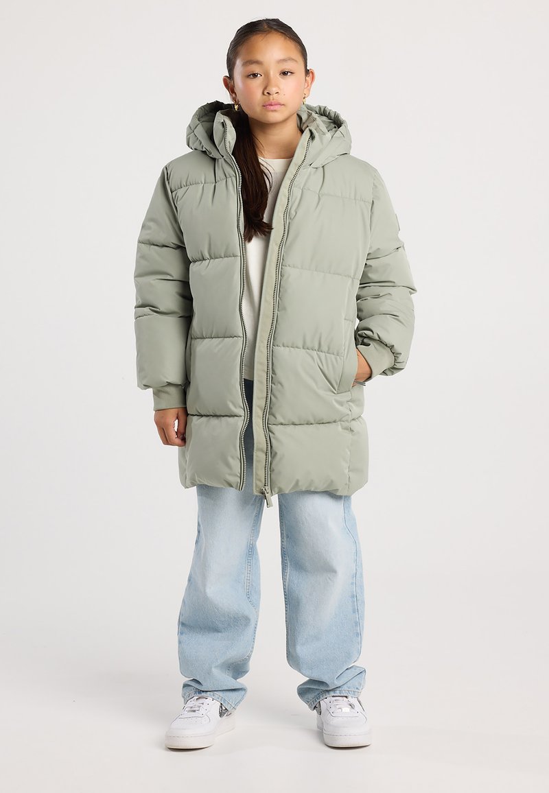 Joven con una chaqueta acolchada con capucha verde claro, jeans azules claros y zapatillas blancas, de pie con las manos en los bolsillos.