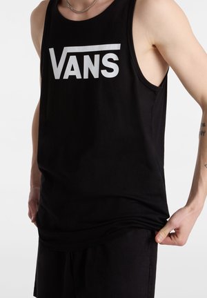 Homme portant un débardeur noir avec le logo blanc "VANS", pantalon noir, chaîne en argent autour du cou, tenant l'ourlet du t-shirt de la main droite.