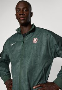 Green zip-up jacket with a textured pattern of letters. Features a white Nike logo and a red lion emblem on the chest.