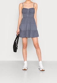 Šedé stupňovité šaty s vyšívaným květinovým motivem, tenkými ramínky a předními knoflíky. Na sobě má bílé tenisky s vysokou podrážkou a drží černou tašku.