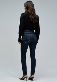 Vaqueros de mezclilla azul oscuro con cintura alta, cuenta con dos bolsillos traseros y pierna ajustada. Combinados con una camiseta negra de manga larga y tacones negros.