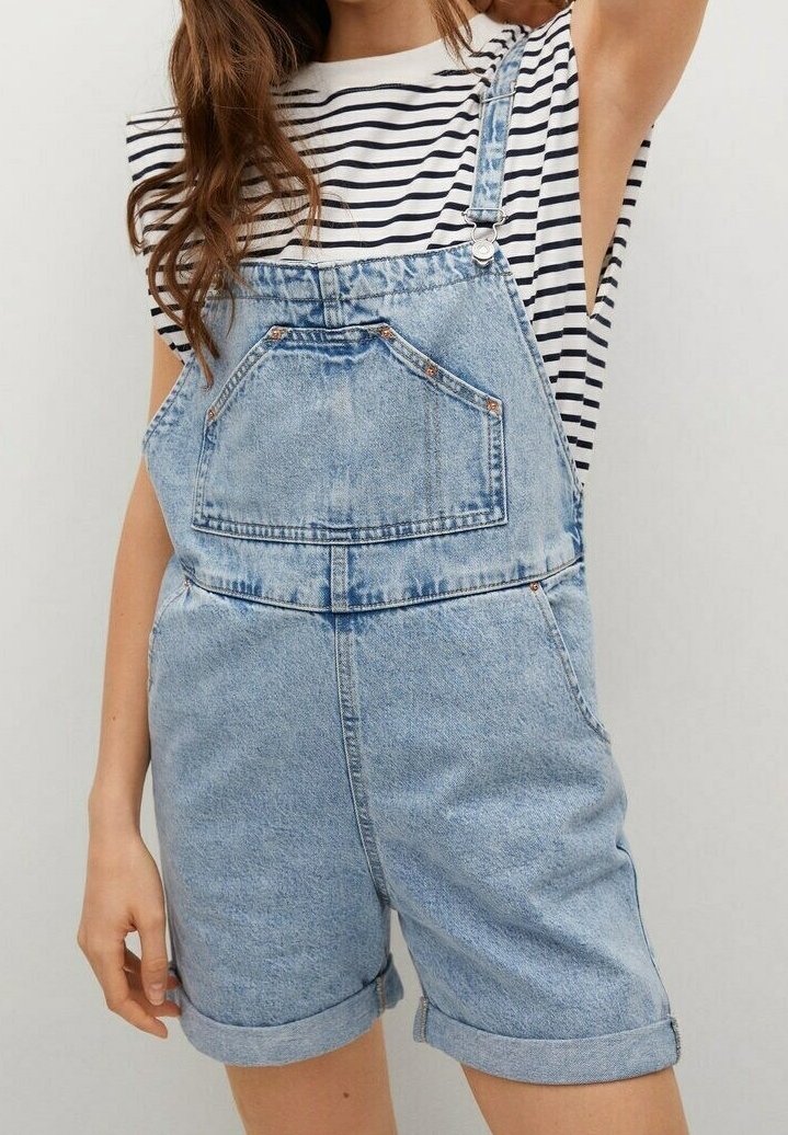 Femme portant une salopette courte en denim bleu clair avec poche frontale et revers roulés, associée à un t-shirt à manches courtes rayé noir et blanc.