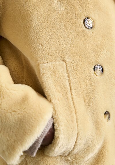 Faux-Fur-Mantel in sanftem Beige, mit doppelter Knopfleiste, zwei sichtbaren Knöpfen und einer seitlichen Tasche mit plüschiger Textur.