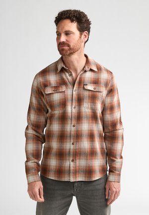 Man met kort krullend haar en baard, draagt een oranje-beige geruit overhemd met lange mouwen en grijze broek, staand tegen een effen achtergrond.