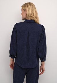 Navy plava bluza s teksturiranim završetkom, s dugmadima na manžetama i nabora na leđima. Krojen podstavljeni stil i mekani materijal. Glatki, opušteni oblik.