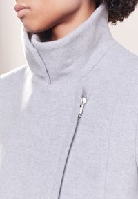 Šedý kabát z vlněné směsi s vysokým límcem, bočním zipem a měkkou texturou, představující čistý, minimalistický design.