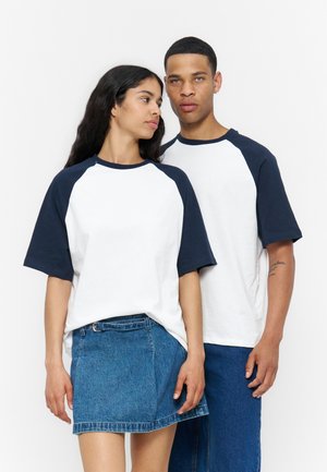 Jeune femme et homme debout côte à côte portant des T-shirts blancs assortis avec des manches bleu marine ; femme en jupe en jean, homme en pantalon en jean.
