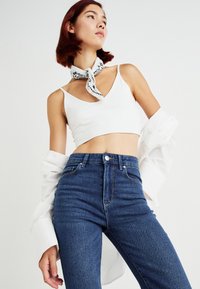 Λευκό κοντό μπλουζάκι συνδυασμένο με ψηλόμεσο σκούρο μπλε τζιν. Ένα κασκόλ με σχέδιο σε μαύρο και άσπρο δεμένο γύρω από το λαιμό. Ανοιχτό φόντο.