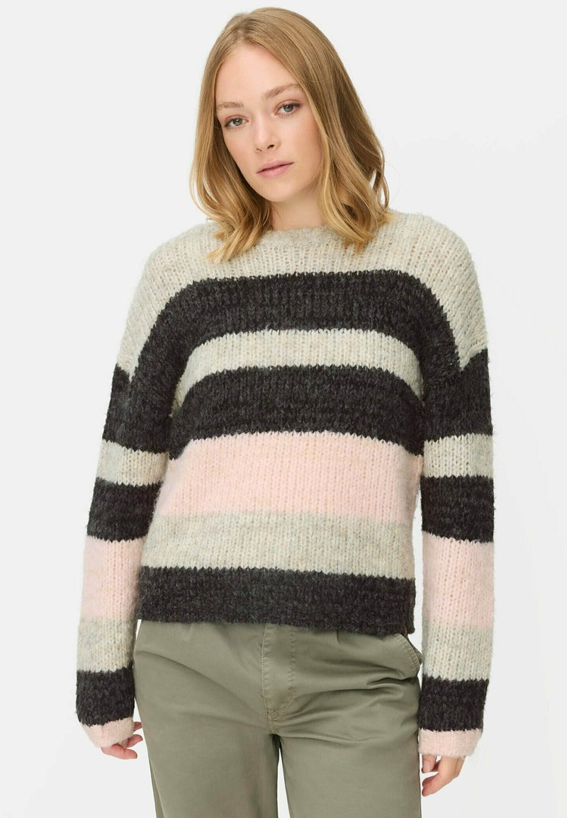 Vrouw in een chunky gebreide trui met brede horizontale strepen in zwart, crème en roze, gecombineerd met olijfgroene broek.