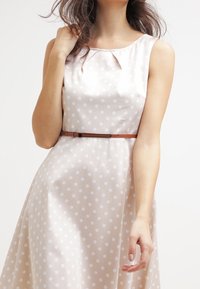 Vestido beige con lunares blancos, diseño sin mangas y corsé ajustado. Presenta un cinturón marrón que acentúa la cintura. Textura de tela suave.