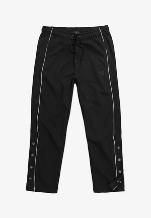 Musta verkkarihousut valkoisella sivuraidalla, kiristysnauhavyötäröllä, painonapeilla ulkosivun lahkeissa ja hillityllä logolla oikeassa reisissä.