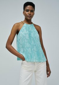 Haut plissé aqua avec motif floral, design à col halter et accents en chaîne dorée aux épaules, associé à un pantalon large de couleur claire.